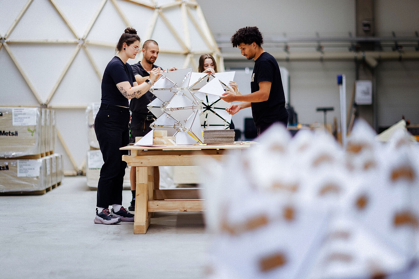 Architekturprojekt am Werktisch Vier Personen arbeiten an einer geometrischen Struktur aus Dreiecken auf einem Holztisch in heller Werkhalle.
