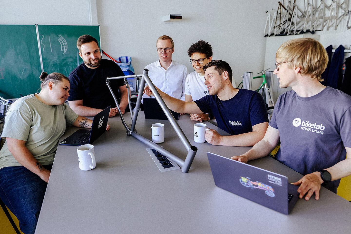 Teamarbeit im Bikelab Sechs Personen sitzen um einen Tisch, betrachten gemeinsam einen Fahrradrahmen, Laptops und Tassen vor sich.