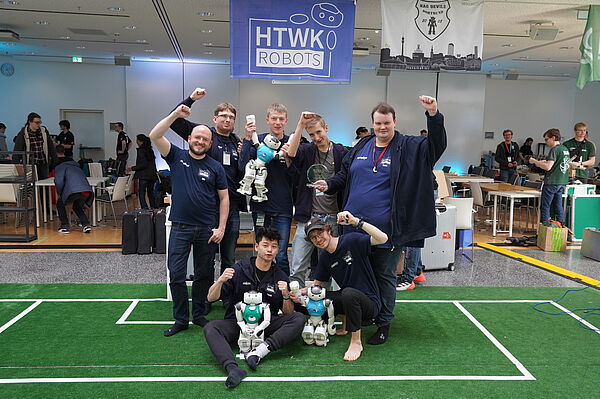 Menschen mit humanoiden Robotern auf kleinem Fußballfeld indoor