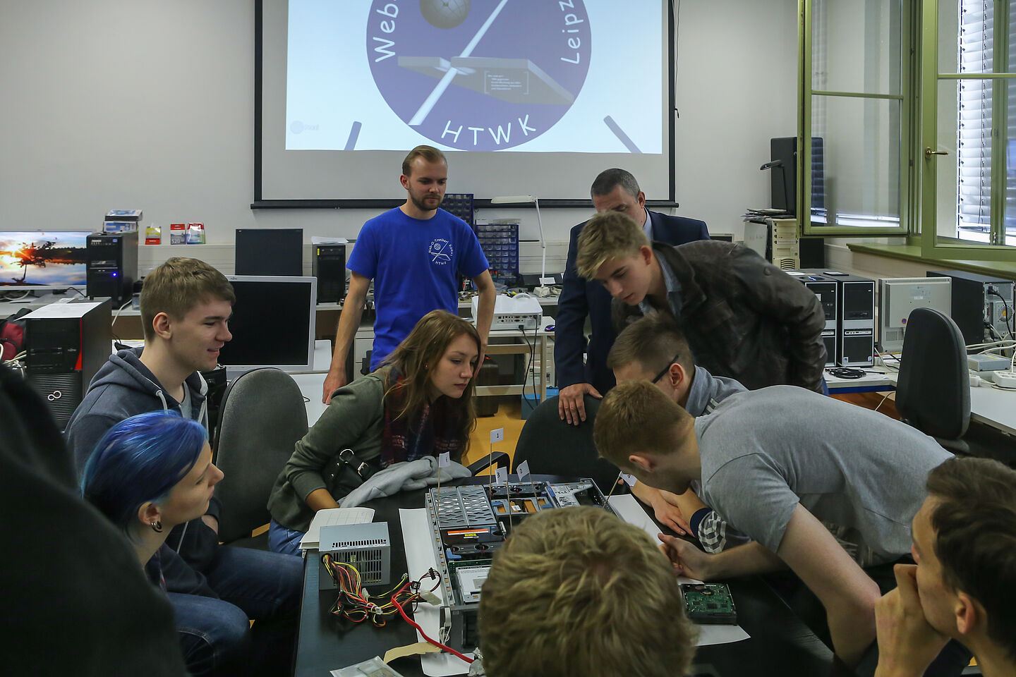 Die studentische Arbeitsgruppe Web-O-Troniker betreut Studierende des 1. Semester der Elektrotechnik und Informationstechnik. Mehrere Studierende sitzen um ein elektronisches Gerät und untersuchen es. Im Hintergrund befindet sich das Logo der Web-O-Troniker auf einem großen Bildschirm.