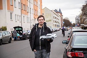 Informatik-Absolvent Andreas Amann steht in einer Leipziger Straße mit einem Modellauto in der Hand