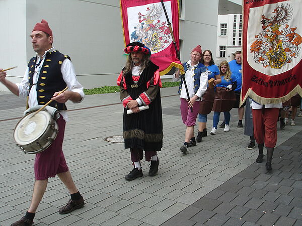 In mittelalterlicher Zunft läuft eine Schlange von Menschen über einen gepflasterten Weg. Viele von ihnen halten große Stoffbanner, auf denen Wappen aufgedruckt sind.