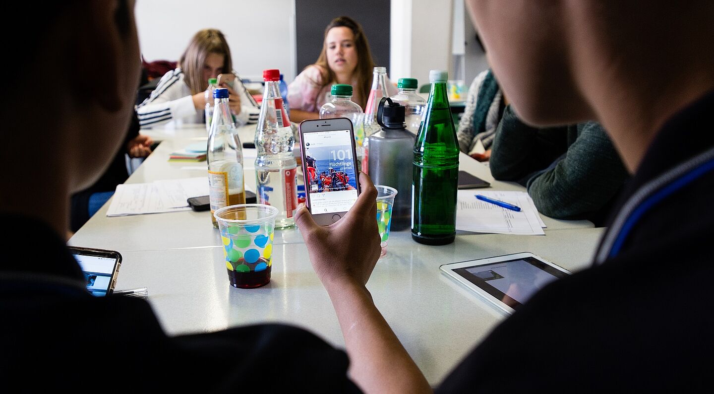 ein Tisch mit mehreren Person, zwei halten ein Smartphone und schauen rein