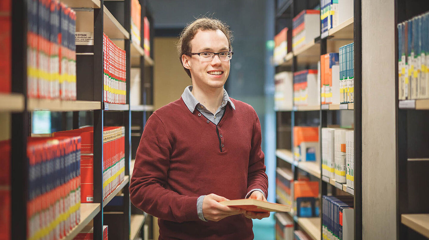 Ein Student zwischen zwei Bücherregalen in der Bibliothek hält ein Buch in der Hand.