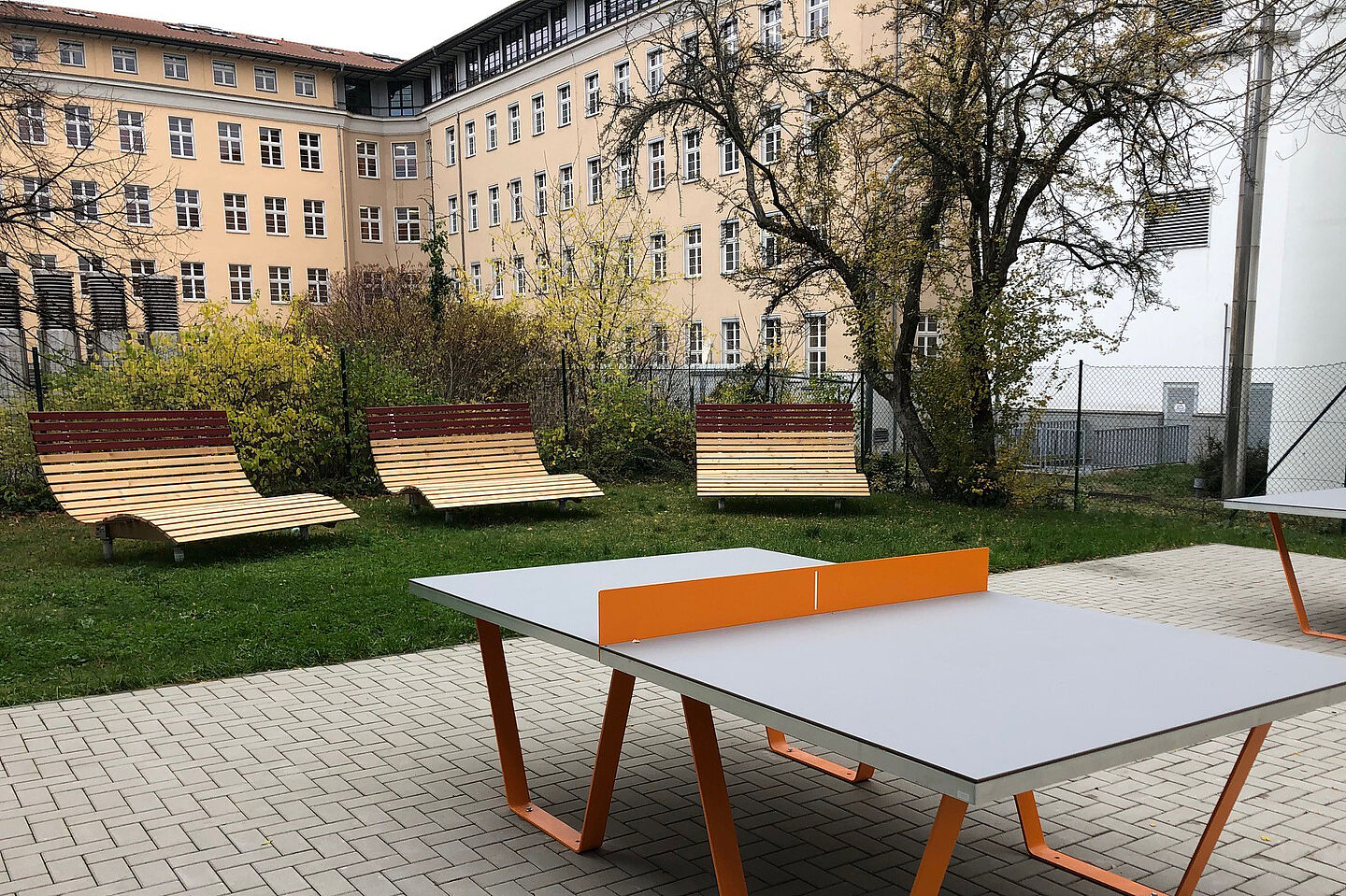 Blick auf eine moderne Tischtennisplatte und drei dahinterstehende Sonnenliegen aus Holz im Innenhof von HTWK-Gebäuden.