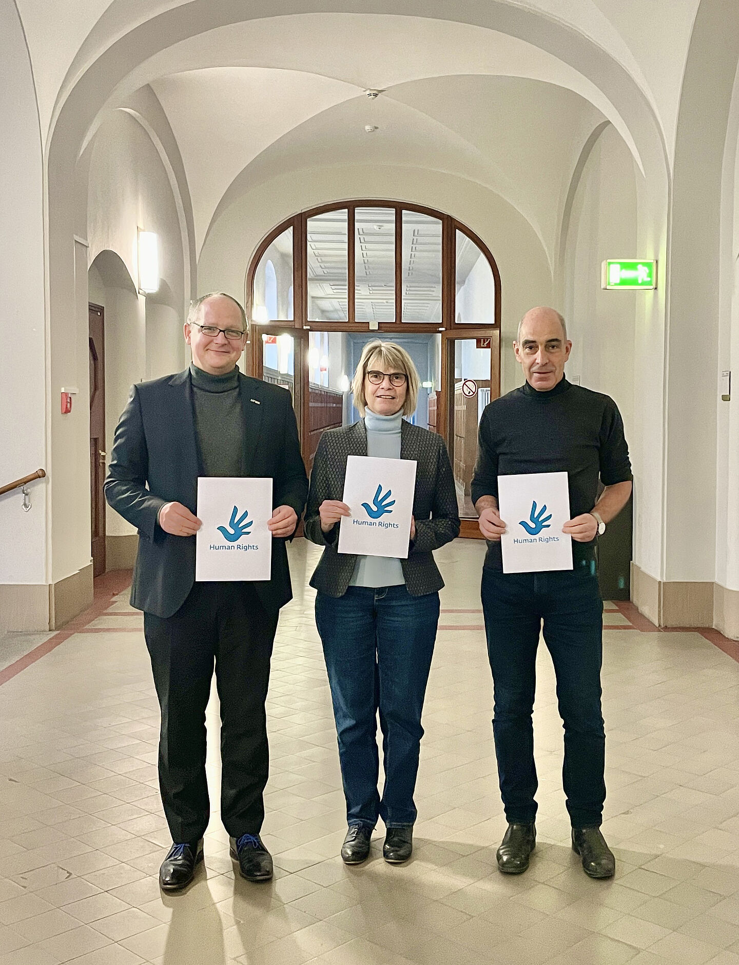 drei Personen mit Schildern in der Hand stehen auf einem langen Flur