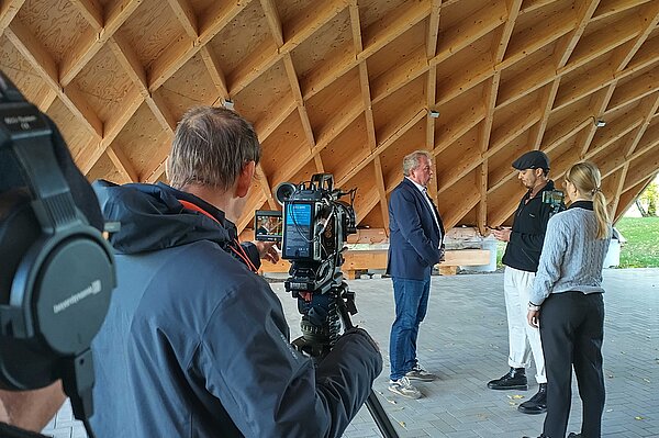 Experte im Interview Menschen stehen für einen Videodreh unter einer Holzdachkonstruktion