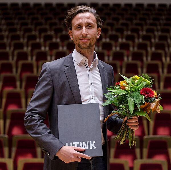 Der Preisträger Max Vollmering hält die Urkunde des Dissertationspreises in der einen Hand und einen Blumenstrauß in der anderen. 