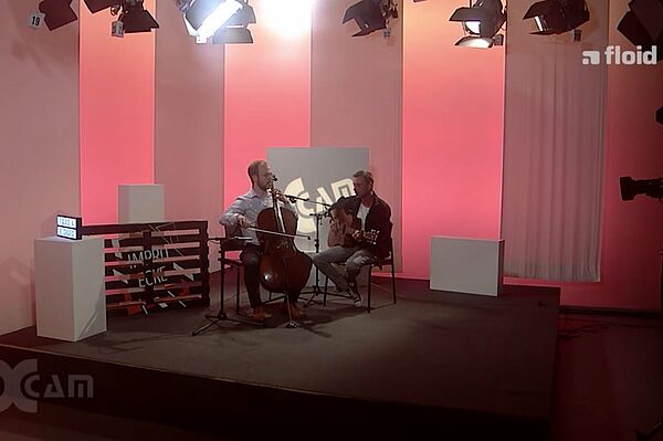 Screenshot eines Filmbeitrags, zwei Musiker mit Cello und Gitarre auf der Studiobühne
