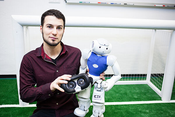 Informatik-Absolvent Marcel Göbe hält einen Nao-Roboter sowie eine VR-Brille in den Händen.