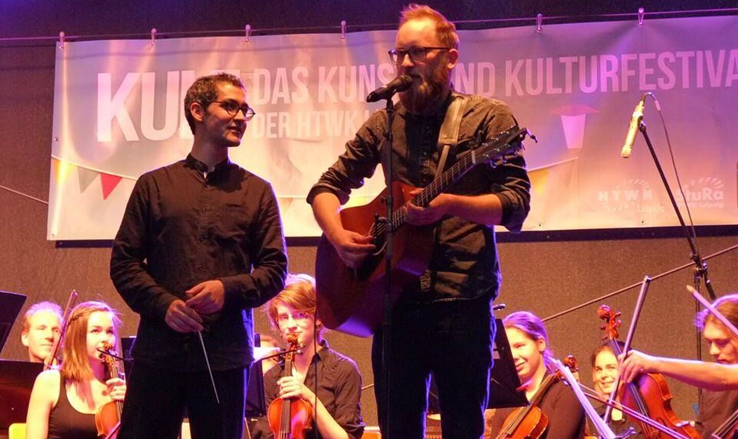 Das HTWK Orchester mit dem Sänger Max von Wegen. Er steht auf der Bühne mit seiner Gitarrre und singt, danaben steht der Dirigent.