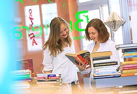 Zwei Studentinnen betrachten gemeinsam ein aufgeschlagenes Buch in der Bibliothek