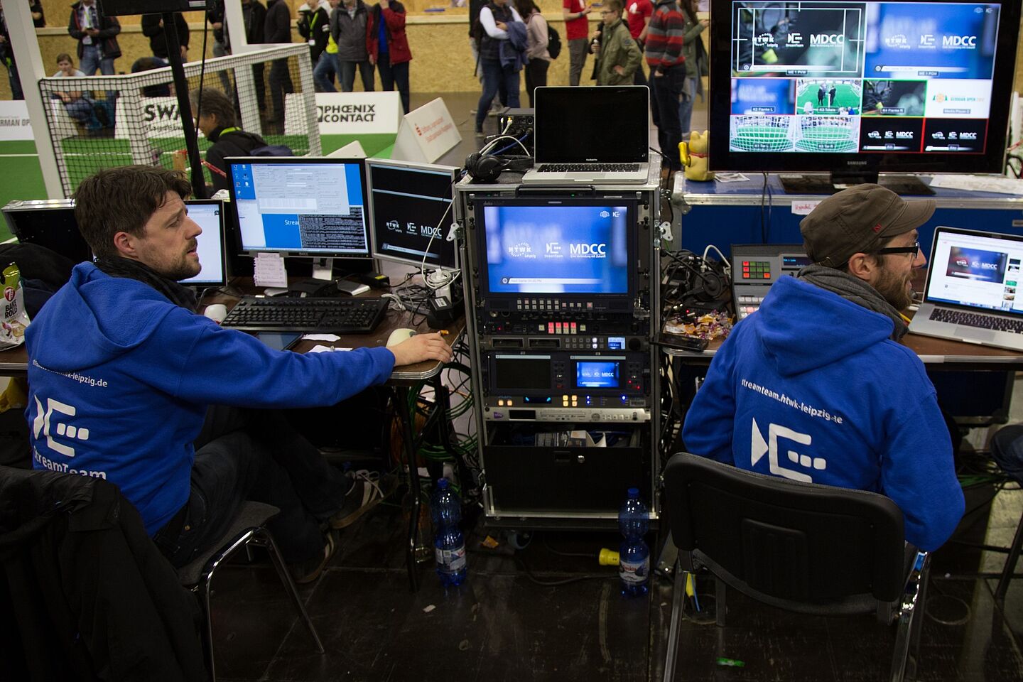 Zwei Studenten bzw. zwei Mitarbeiter der HTWK sitzen vor mehreren Bildschirmen und streamen ein Sportevent. 