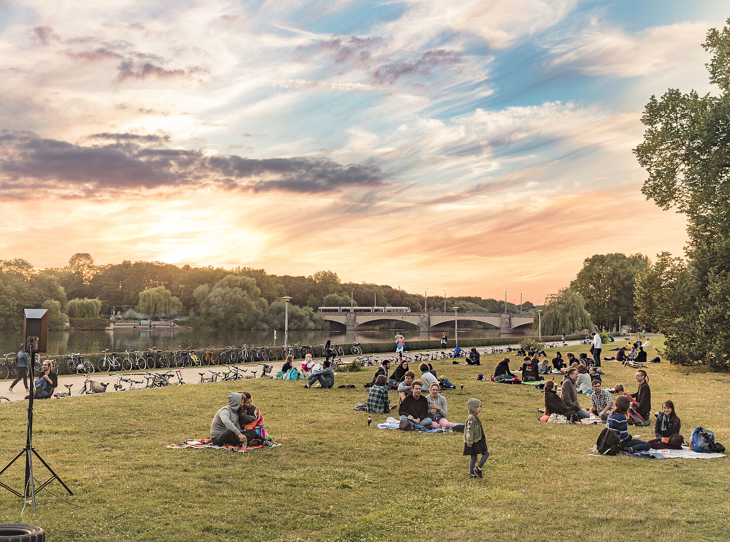 Menschen sitzen in einem Park an der Leipziger Elster. Der Himmel ist rot-bläulich.