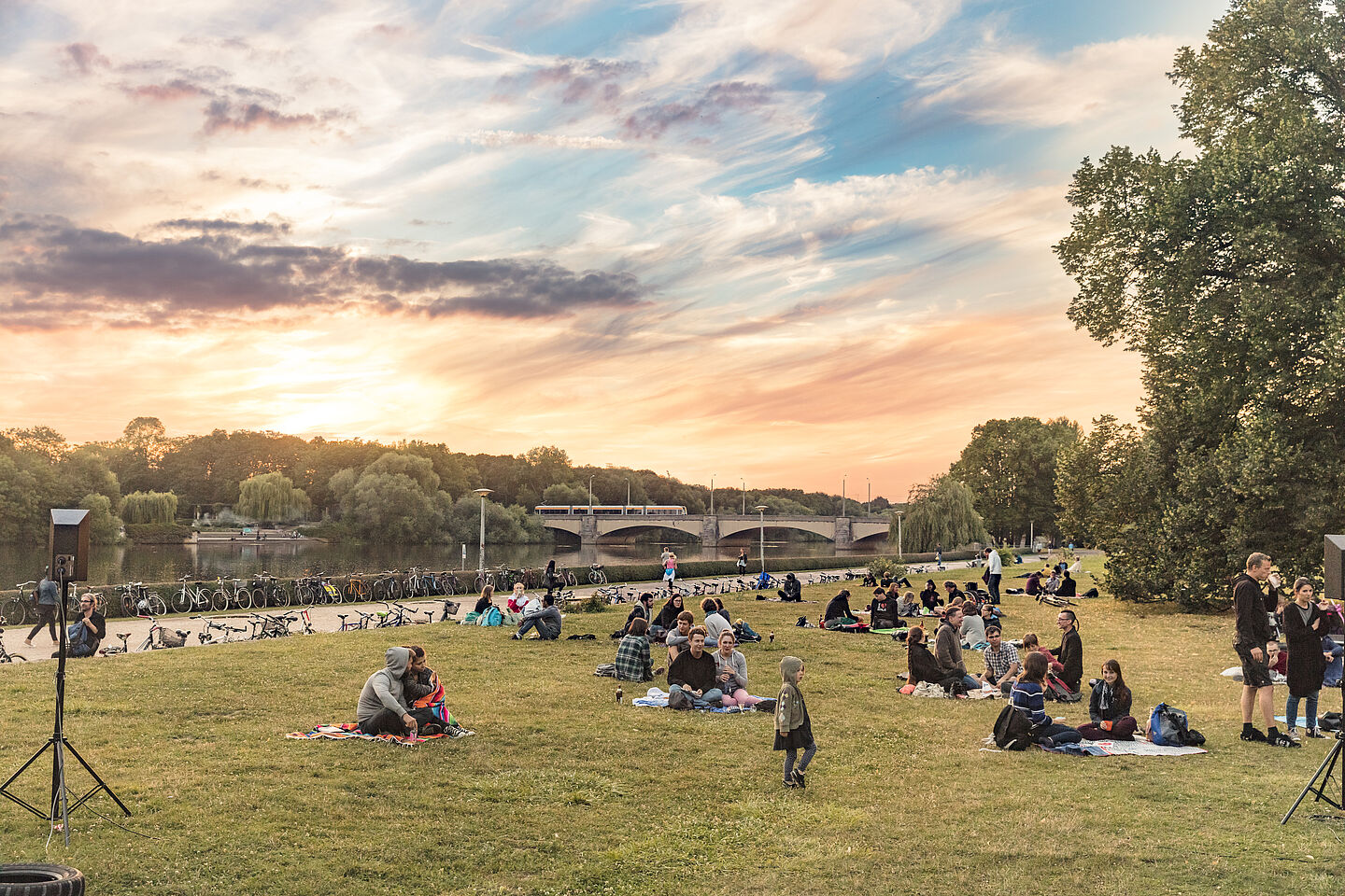 Menschen sitzen in einem Park an der Leipziger Elster. Der Himmel ist rot-bläulich.