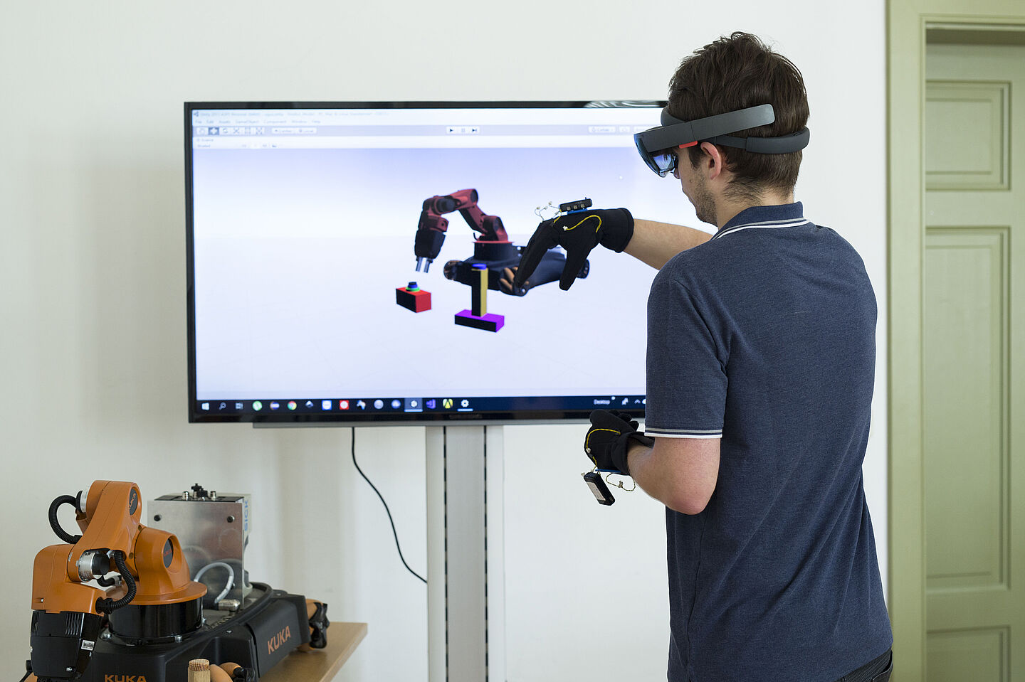 Person mit VR-Brille und Joysticks bewegt in der virtuellen Realität Roboterteile. Das Ergebnis wird auf einem Bildschirm gezeigt.