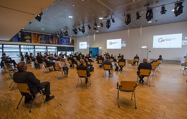 Saal mit locker verteiltem Publlikum auf Stühlen.