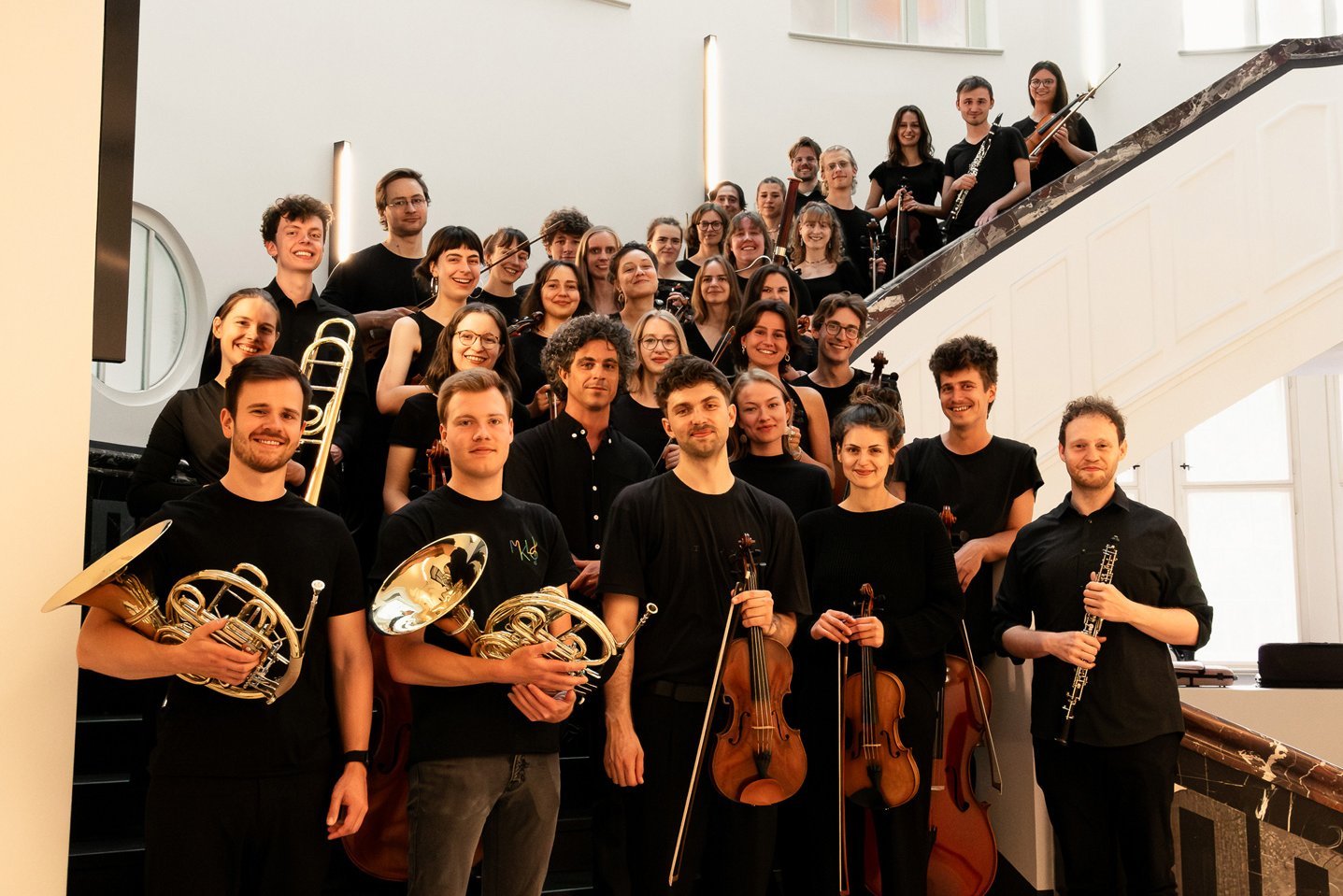 HTWK Orchester Leipzig SoSe25 Gruppenbild des Orchesters mit ihren Instrumenten auf einer Treppe in einem HTWK-Gebäude.