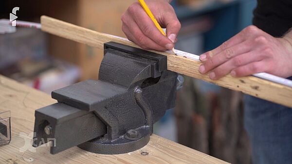 Schraubstock, der ein Stück Holz einfasst. Jemand misst das Stück Holz und markiert mit Bleistift