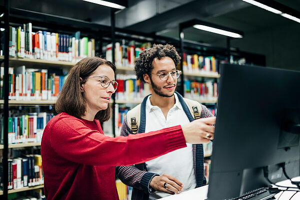 Eine Bibliotheksmitarbeiterin zeigt einem Studenten etwas am PC.