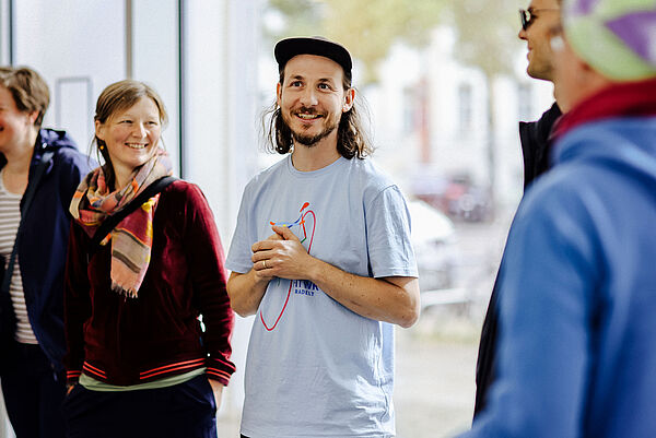 Ein Mann steht in der Mitte eines Raumes und lächelt. Er trägt ein hellblaues T-Shirt und eine schwarze Kappe. Der Mann hat schulterlanges, braunes Haar und einen Bart. Er hält seine Hände vor sich zusammen. Neben ihm, links im Bild, steht eine Frau, die ebenfalls lächelt. Sie trägt eine bunt gemusterte Schals und eine rote Jacke. Im Hintergrund sind unscharf weitere Personen und die Fensterfront eines Gebäudes zu sehen. Die Szene wirkt freundlich und einladend.