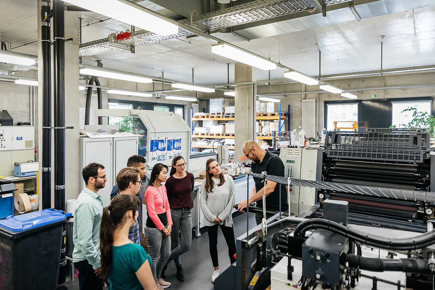 Kleine Personengruppe steht im großgeräumigen Drucksaal vor einer Druckmaschine