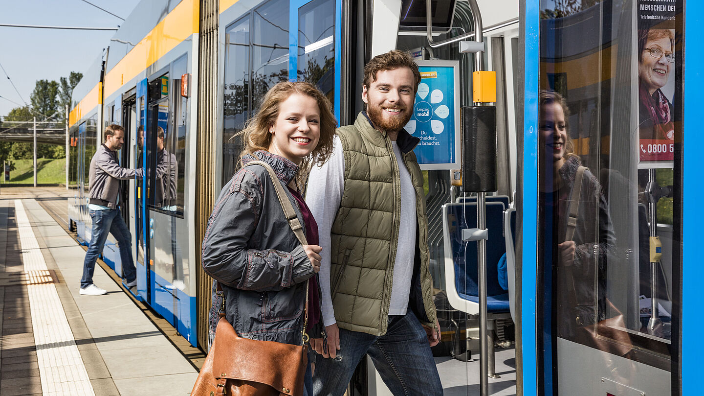 Zwei Menschen steigen in eine Leipziger Straßenbahn ein. Bei schauen lächelnd in die Kamera.