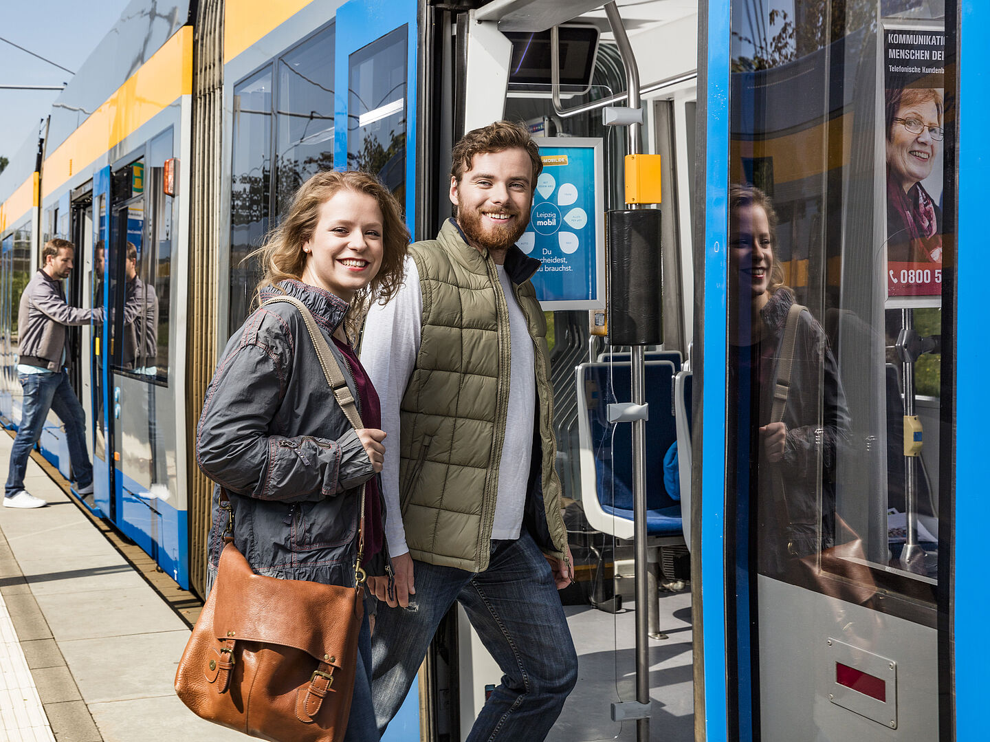 Zwei Menschen steigen in eine Leipziger Straßenbahn ein. Bei schauen lächelnd in die Kamera.