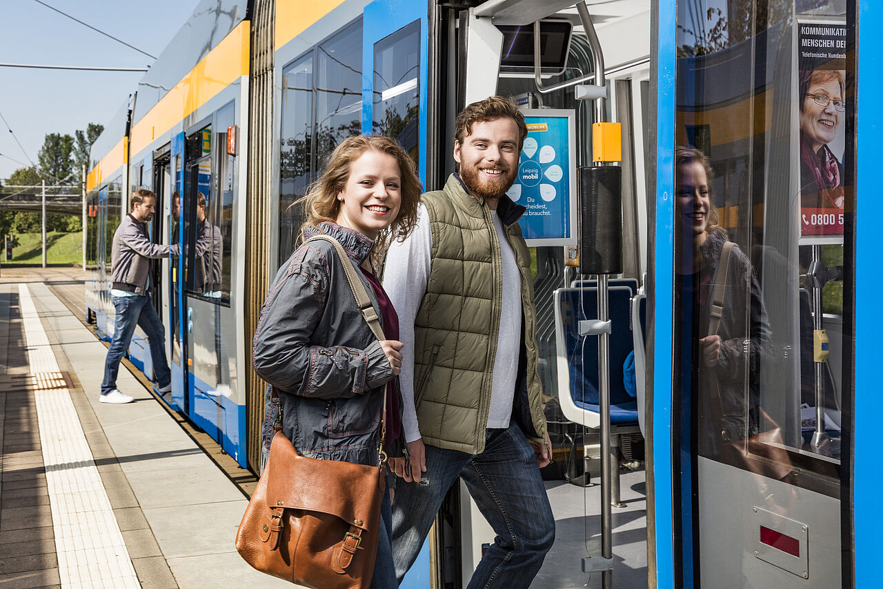 Zwei Menschen steigen in eine Leipziger Straßenbahn ein. Bei schauen lächelnd in die Kamera.