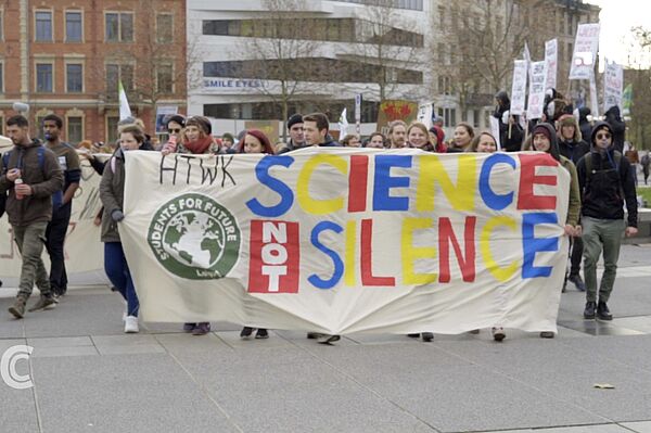 Screenshot der Campus Compact-Sendung zur Klimastreikwoche: Menschen laufen auf der Straße und ragen ein Soffplakat mit der Auschrift „HTWK Students for future Science not silence“ 
