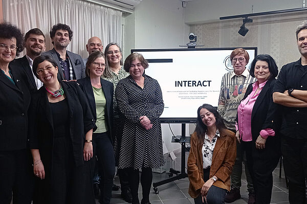 Das Foto zeigt 12 Frauen und Männer stehend vor einem Bildschirm mit der Aufschrift INTERACT