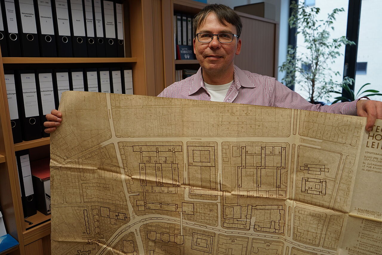 HTWK-Archivar Benjamin Schäf steht in seinem Büro und präsentiert eine riesige vergilbte Karte mit Bauskizzen aus dem Jahr 1970.