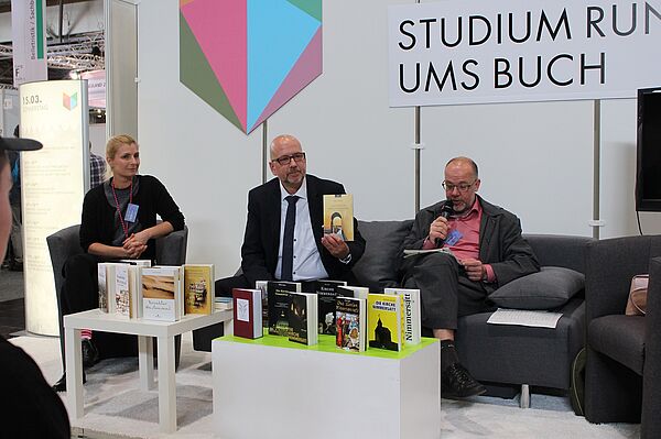 Eine Frau und zwei Männer sitzen auf einer Couch, der mittlere hält ein Buch hoch