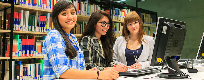 Drei Studentinnen stehen vor dem Computer in der Bibliothek und suchen Informationen über beispielsweise vorhandene Bücher.