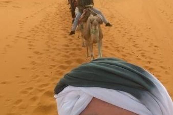 Sebastian Ehrhart reitet in der Wüste auf einem Kamel und schießt das Foto während des Ritts von sich selbst. Im Hintergrund sieht man eine Karawane aus weiteren Reitern.