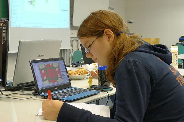 Eine Studentin berechnet etwas auf einem Blatt Papier, neben ihr ist ein Netbook mit dem betreffenden Spiel offen