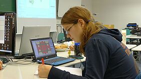 Eine Studentin berechnet etwas auf einem Blatt Papier, neben ihr ist ein Netbook mit dem betreffenden Spiel offen