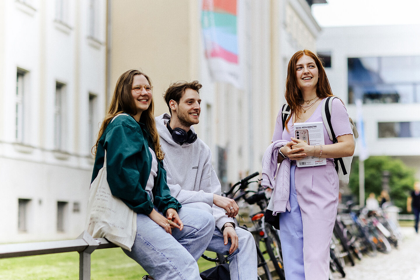 3 lächelnde Studierende vor einem HTWK-Gebäude mit Regenbogenflagge