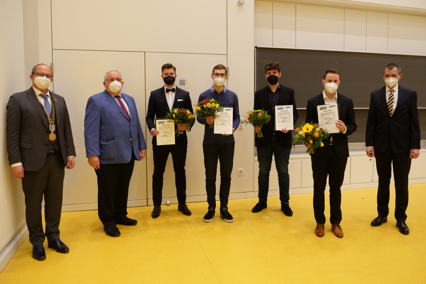 Preisträger stehen mit Urkunde und Blumen in der Hand und mit Maske nebeneinander