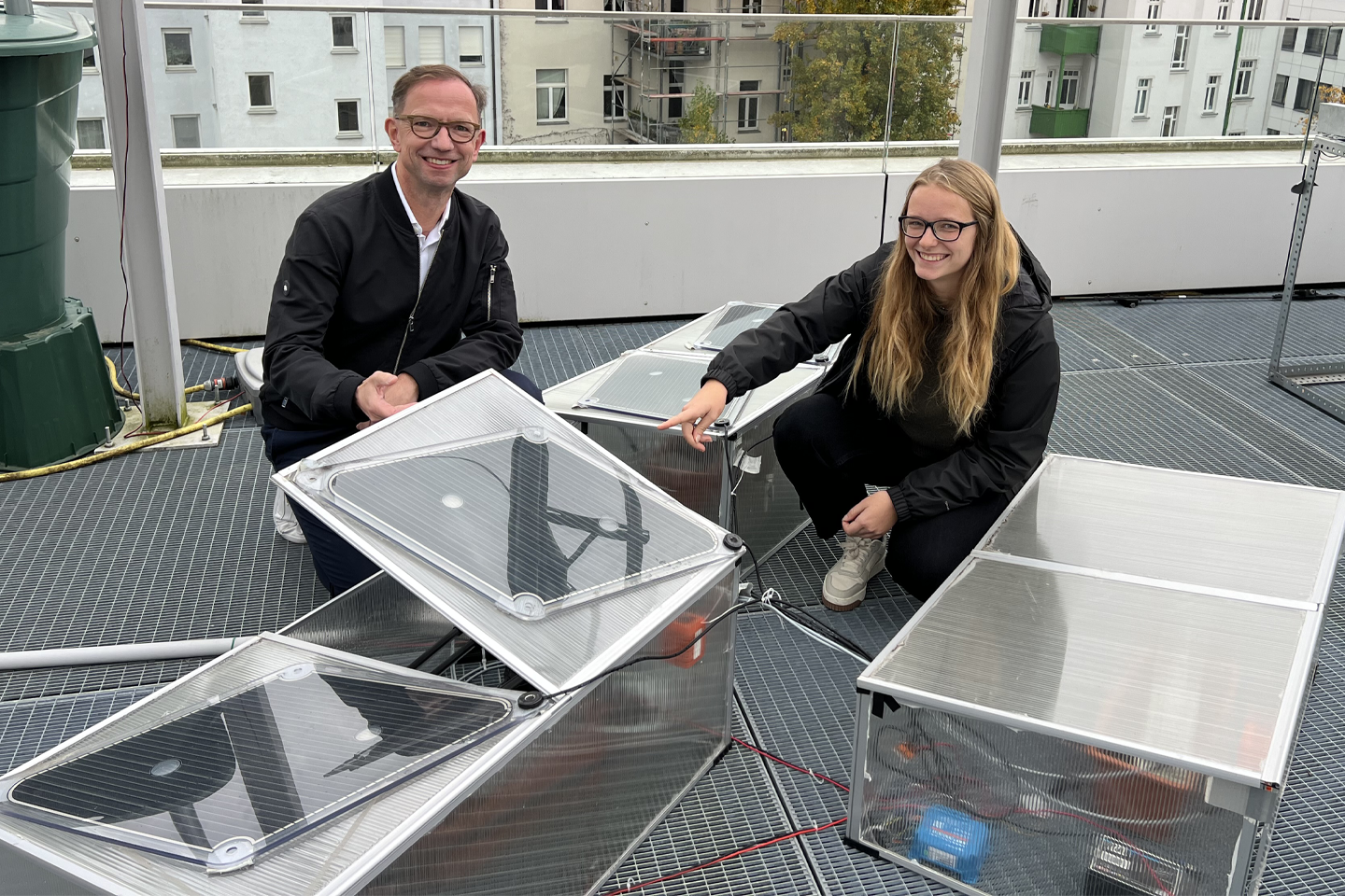 Prof. Rudolph und Laura hocken vor ihrem Versuchsaufbau mit Mini-Photovoltaikpanels auf dem Dach der Fakultät und blicken in die Kamera