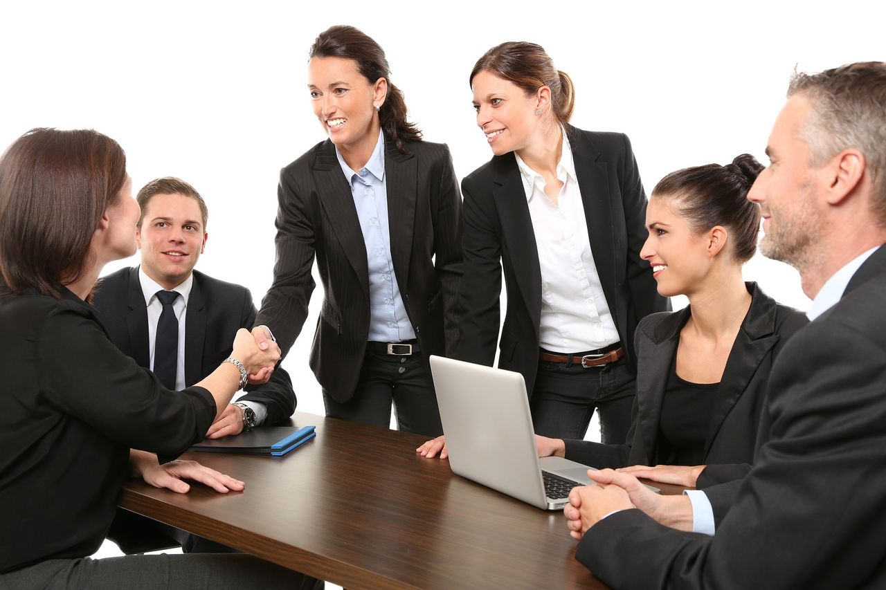 Six people in business suits meet at a table, four women and two men. Two of the women shake hands.