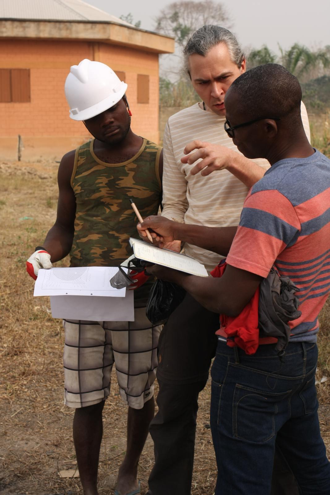 Drei Personen, in der Mitte ein deutscher Ingenieur, umgeben von zwei Dorfbewohnern, unterhalten sich anhand eines Dokuments