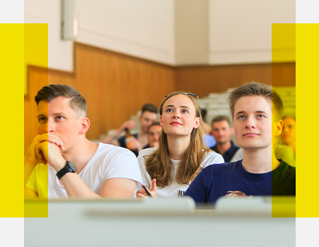 Im Vordergrund sieht man zwei Studenten und eine Studentin im Hörsaal, die gespannt einer Vorlesung lauschen.