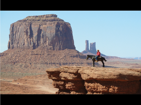 Cowboy auf Pferd steht alleine auf einem Felsvorsprung, im Hintergrund ragt ein rechteckiges Felsmassiv in den Himmel.