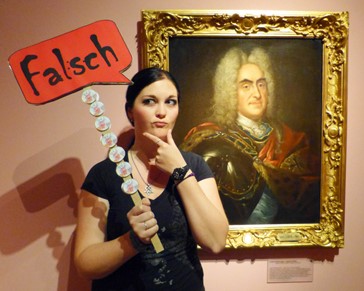 Gaumnitz Franziska Eine Absolventin des Studiengangs Museologie an der HTWK Leipzig steht in einer Ausstellung neben einem Gemälde von August dem Starken. Sie hält ein Schild mit der Aufschrit: „Falsch“, hoch. Sie greift sich nachdenklich ans Kinn.