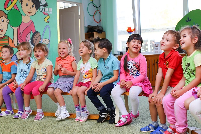 Mehrere kleine Kinder sitzen in einem Kindergarten lachend auf einer Bank nebeneinander.